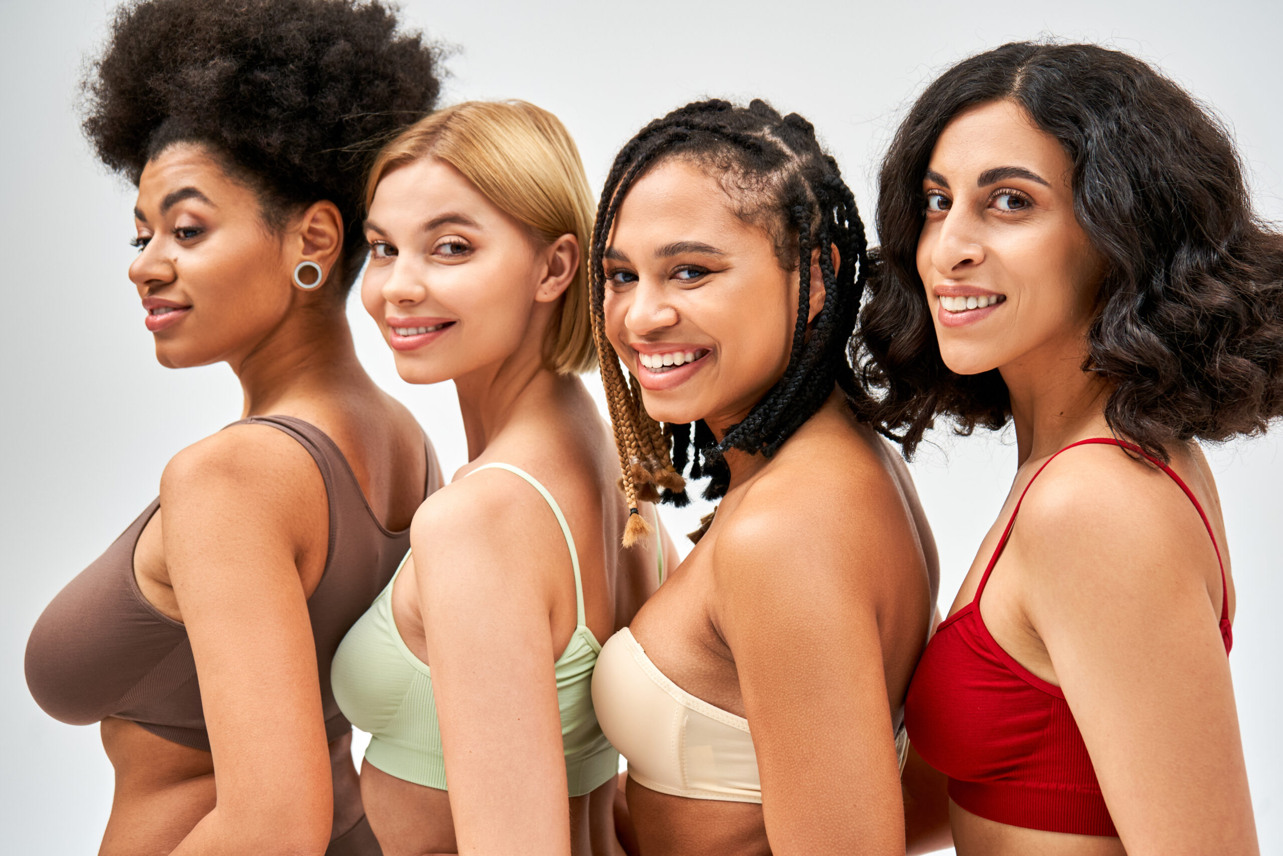 Portrait of cheerful and multiethnic women in colorful bras looking at camera while posing together isolated on grey, different body types and self-acceptance concept, multicultural models